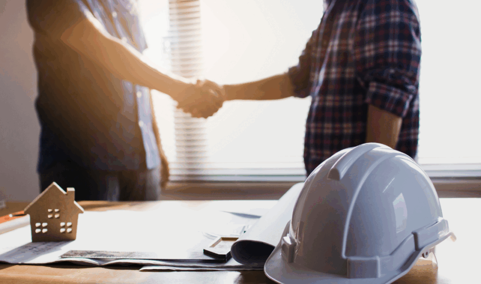 Contractor shaking hands over construction plans with a hard hat on the table, symbolizing agreement and contract in Colorado construction.